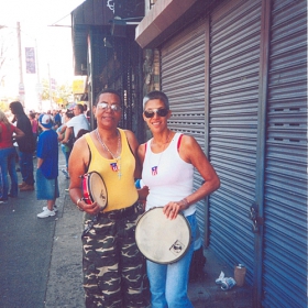 Photo of two women wearing tank tops and jeans standing on a street holding drums.