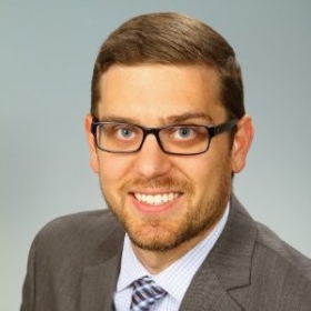 Steven Malick, a man smiling wearing a suit and striped tie and black rectangular glasses. 