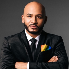 Julio Roman, who has a beard and is wearing a suit and tie with a rainbow pocket square, crosses his arms and looks into the camera looking serious.