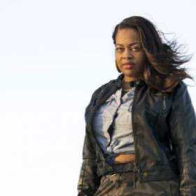Venice brown, wearing a gray shirt and jacket, stands looking seriously at the camera. Her long black hair blows behind her in the wind.