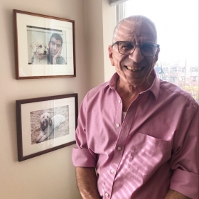 Joseph Canarelli, a balding man with glasses, wearing a pink shirt, stands in front of a window. There are photos of a dog and a man on the wall next to him.