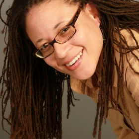 Burley Tuggle, who has long hair and rectangular glasses, is leaning over and smiling at the camera.