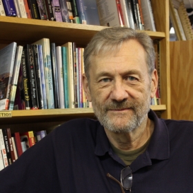 Michael Bronski, wearing a black shirt, poses in front of a bookshelf.