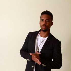 Brian Epps, wearing a black jacket, white shirt, and gold necklace, looks seriously at the camera. He holds his hands together. 