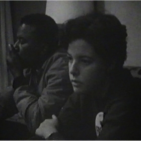 Photo of Carol Glassman, a young woman with short dark hair wearing a button on her chest. She has a serious expression.