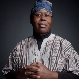 James Credle, a Black man wearing a blue head covering and striped shirt, looks seriously at the camera. His hands are folded. He is wearing glasses.