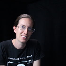 Emma Wilcox, a woman with her hair pulled back and rectangular glasses, is wearing a black shirt and posed in front of a black background.