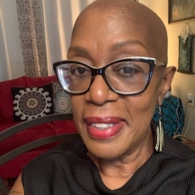 Erica Harrison, a Black woman with a shaved head a large Black glasses. She is wearing earrings and a black shirt and is seated in a living room with a couch in the background.