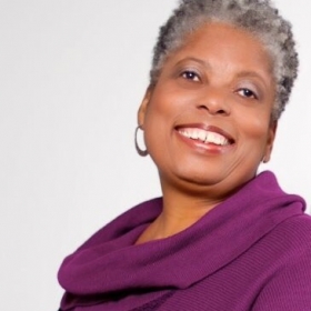 Janyce Jackson Jones, a woman with short gray hair, smiles in front of a white background. She is wearing a magenta scarf and earrings.