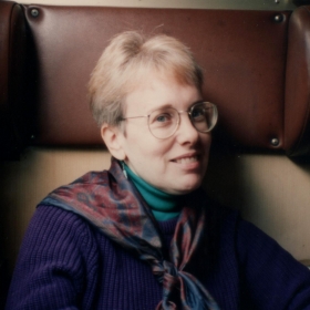 Gail Malmgreen, a woman with round white glasses, a green turtleneck, and a purple scarf and sweater, and short blond hair.
