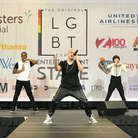 Lovari while performing on a stage with a rainbow logo and several corporate logos, with two backup dancers.