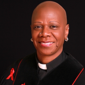Jerri Mitchell-Lee, a woman smiling with a shaved head, earrings, wearing a reverend's collar and a red ribbon on her chest. 