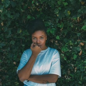 Jasmine Mans against a background of leaves. Her hand is on her chin. She is wearing dark turquoise ppants and a lighter turquoise shirt.