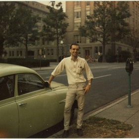 John, a young man, stands in front of a car wearing tan pants and a white shirt. it is an old photo.