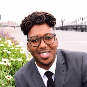 Kareem, wearing a suit and tie, posed outdoors by a flowering plant and a river. He has round glasses and an elaborate hairstyle.