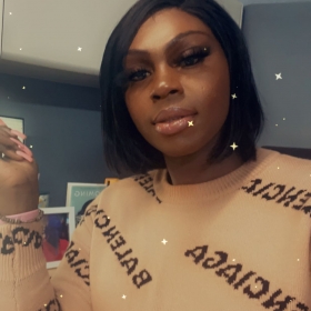 Maya Williams, a woman wearing a tan Balenciaga shirt, poses with her hand to her side. She is wearing glasses and has black shoulder length hair.