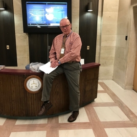 Bert Mrozik, posing on a desk in a government building. He is wearing dress pants, a dress shirt, a tie, and black rectangular glasses, and he is bald.