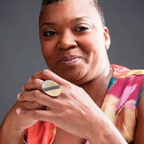 June Dowell Burton, wearing a brightly colored shirt, smiles at the camera with her hands folded. She is wearing a large circular ring and has short hair.