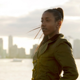 Patreese Johnson, a woman with a ponytail, is wearing a green shirt and standing in front of a blurred city skyline.