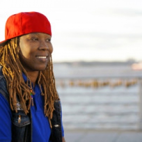 Renata Hill, a Black woman wearing a blue shirt, black best, and red baseball cap turned backwards, smiles against a blurred outdoor background.