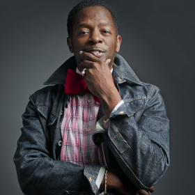Rodney Gilbert, wearing a plaid shirt and a black jacket, poses with his hand on his chin.