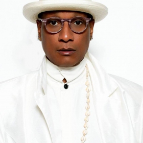 Sir Dane, wearing round dark glasses, two gold necklaces, a white hat, a white blazer, and a white shirt.