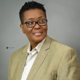 Jacquelyn Hollard, a woman with short hair and rectangular black glasses, smiles in front of a gray background. She is wearing a brown blazer and white button down shirt.