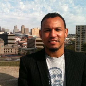 Perris Straughter, wearing a black jacket and a white shirt featuring a man's face, smiles at the camera. He is pictured against a city skyline.