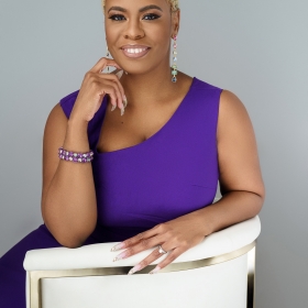 Dana Davis, wearing a purple tank top, sits in a white chair against a gray background smiling at the camera.