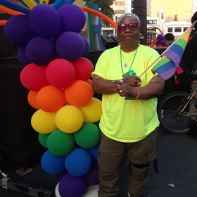 Tiney, wearing a bright green shirt and a backward baseball cap, poses by a stack of rainbow balloons at a pride parade.