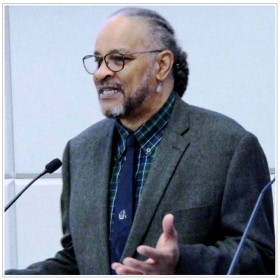 Gary Paul Wright, a man with a ponytail and glasses, wearing a suit, stands in front of a microphone, gesturing.