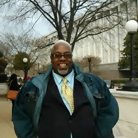 Aaron Frazier stands outdoors in front of a tree. He is smiling and wearing a suit and winter coat.