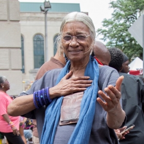 Amina Baraka, an older woman with gray hair pulled back, wearing a gray shirt and a blue scarf, stands with her right hand on her heart and her left hand outstretched toward the camera.