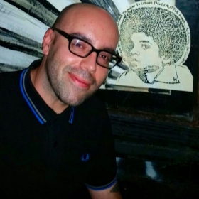 Aleix Martinez, smiling at the camera. He is bald and wearing a black shirt. He is posing in front of an art piece of a young Black person with an Afro.