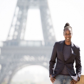 Anita Dickens, who has her hair in a bun and is wearing jeans, a blouse, and a blazer, poses in front of the Eiffel Tower.