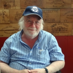 Bob Cartwright, an older man with white hair and a beard, sits in a chair. He is wearing a blue shirt and baseball cap and is seated in front of a brown wooden wall with carvings.
