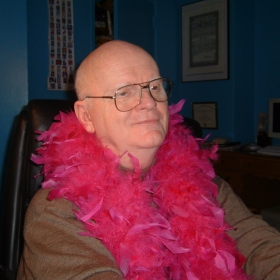 Louie Crew Clay, a balding older man with glasses and a pink feather boa, is seated indoors.