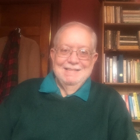 Bill Courson, a balding older man with round glasses wearing a green sweater and turquoise collar, smiles at the camera. He is seated in front of a bookshelf.