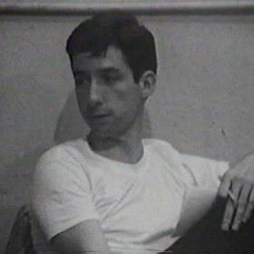 Tom Hayden, a young man with dark hair with a serious expression and wearing a white t-shirt.