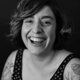 Marina Carreira, who has shoulder length wavy hair, a tank top, and tattoos on her arms, looks at the camera and looks as though she is laughing. The photo looks modern but is in black and white.