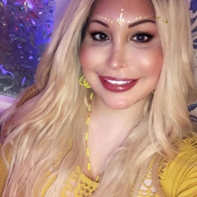Juliana Moores, smiling at the camera wearing a yellow shirt. She has long blond hair and is waering bright sparkly makeup.
