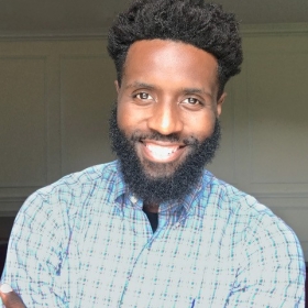 Larry Lyons, a Black man with a beard wearing a checkered shirt, smiling.