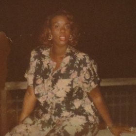Pucci Revlon, wearing a floral dress and earrings, looks into the camera. She appears to be outside. The photo is old.