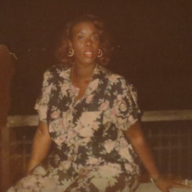 Pucci Revlon, wearing a floral dress and earrings, looks into the camera. She appears to be outside. The photo is old.