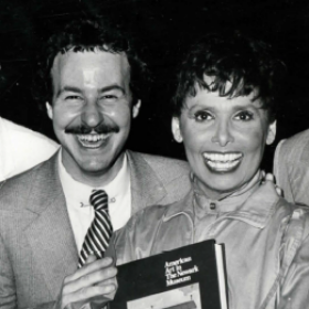 Walter Newkirk, a man with dark hair and a mustache, is wearing a suit and tie and smiling at the camera. He is posing with a woman who is holding a book. The photo is black and white.