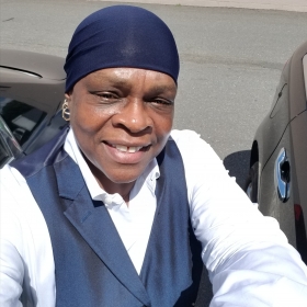 Peggie Miller, smiling and wearing a blue head covering, a white shirt, and a navy vest.