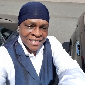 Peggie Miller, smiling and wearing a blue head covering, a white shirt, and a navy vest. 