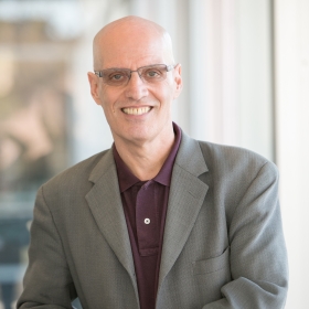 Peter Savastano, wearing a gray blazer and dark purple button down shirt, smiles at the camera. He is bald and is wearing glasses.
