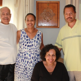 The Proctor Family: a standing man wearing a white shirt, a standing woman wearing blue and white, a seated woman wearing black, and a standing man wearing yellow, posing inside a home.