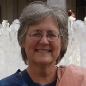 Terri Seuss poses in front of a fountain. She is an older woman with short gray hair and glasses. She is wearing a blue shirt with a pink jacket over her shoulder.